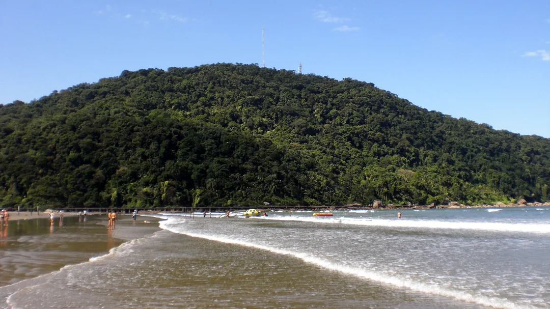 Ondas chegando à areia da Praia do Canto do Forte, com o morro coberto de mata ao fundo, em Praia Grande (SP)