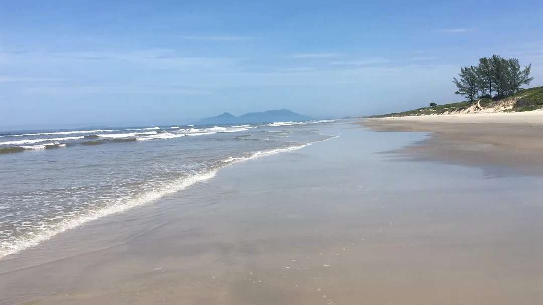 Areia clara e ondas na praia de Ilha Comprida, com morros no horizonte, em Ilha Comprida (SP)
