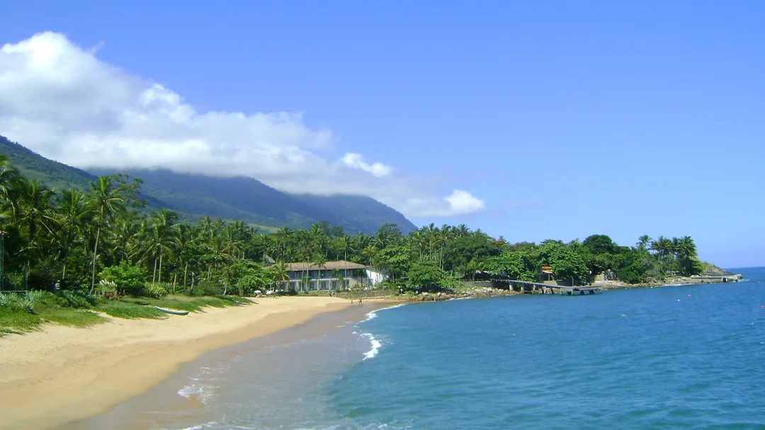 Areia e mar azul da Praia da Feiticeira, com a mata e as montanhas ao fundo, em Ilhabela (SP)