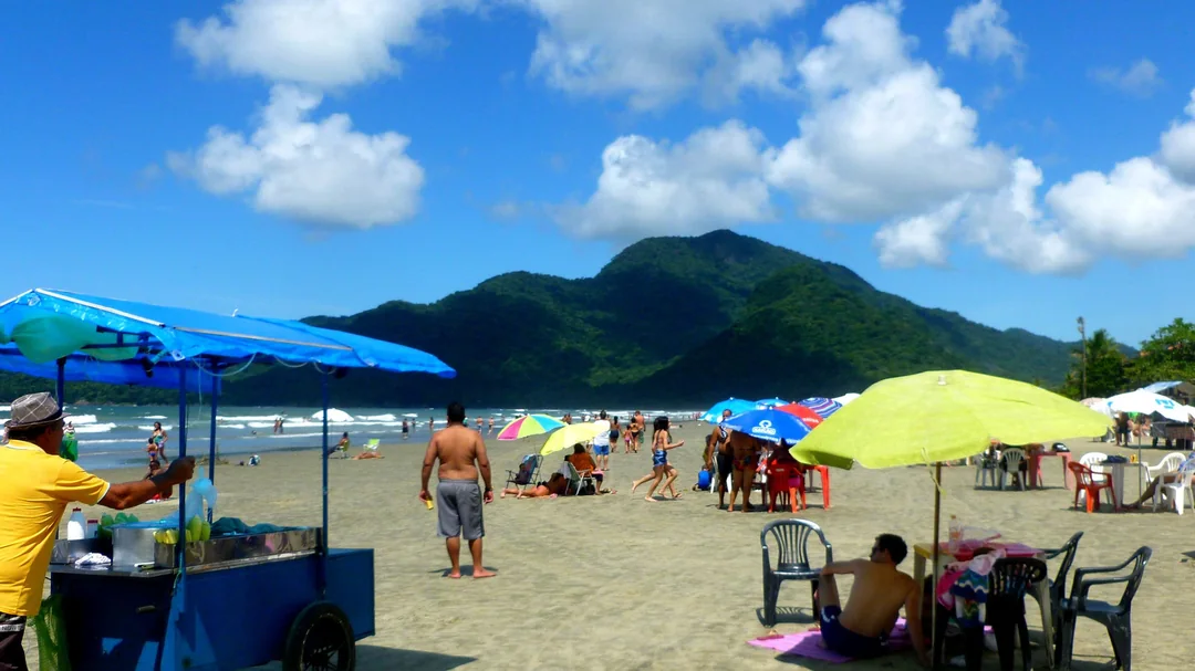 Barracas, guarda-sóis e banhistas na areia da Praia do Guaraú, com morros cobertos de mata ao fundo, em Peruíbe (SP)