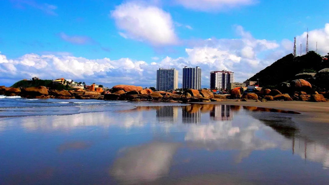 Maré baixa refletindo o céu entre as pedras da Praia do Sonho, com prédios ao fundo, em Itanhaém (SP)