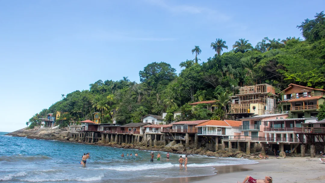 Areia e mar calmo da Praia de Sorocotuba, com casas entre a mata, em Guarujá (SP)
